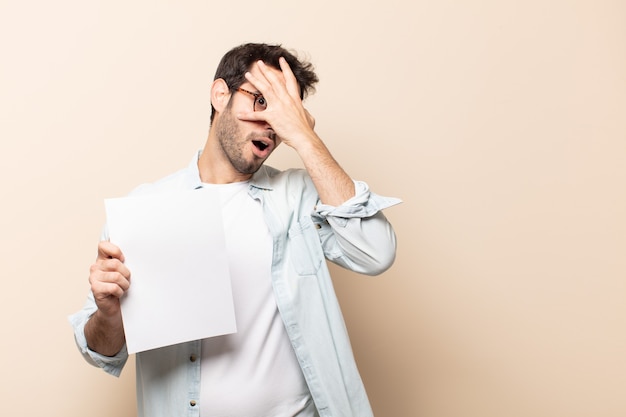 Jeune bel homme ayant l'air choqué, effrayé ou terrifié, couvrant le visage avec la main et regardant entre les doigts