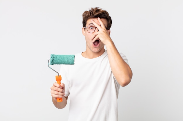 Jeune bel homme ayant l'air choqué, effrayé ou terrifié, couvrant le visage avec la main et tenant un rouleau de peinture