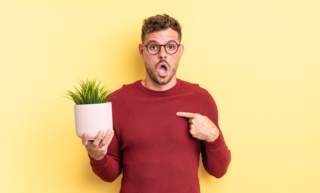 jeune bel homme ayant l'air choqué et surpris avec la bouche grande ouverte, pointant vers lui-même. concept de plante décorative