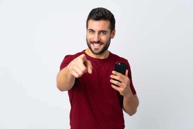 Jeune bel homme avec barbe à l'aide de téléphone mobile isolé sur blanc pointe du doigt vous avec une expression confiante