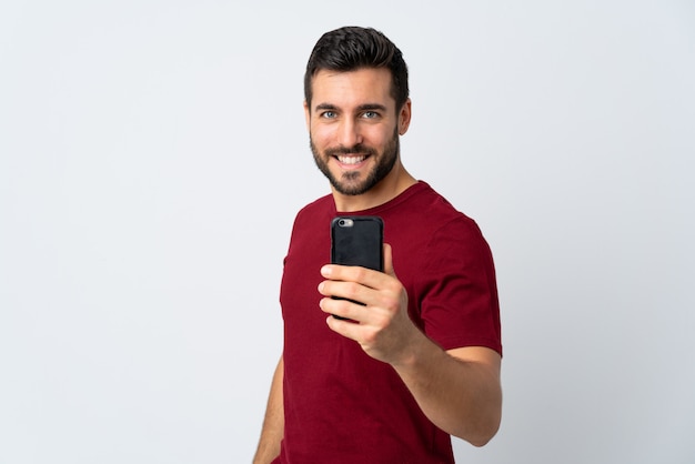 Jeune bel homme avec barbe à l'aide de téléphone portable isolé sur mur blanc avec une expression heureuse