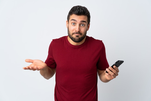 Jeune bel homme avec barbe à l'aide de téléphone portable isolé sur mur blanc faisant des doutes geste tout en soulevant les épaules