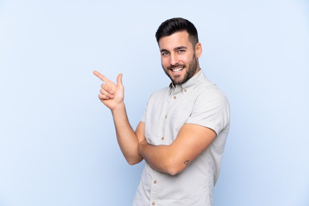 Jeune bel homme avec barbe sur fond bleu isolé pointant le doigt sur le côté