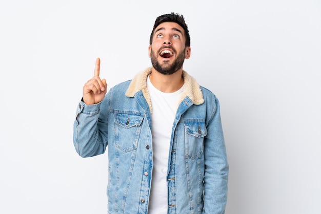 Jeune bel homme avec barbe isolé sur mur blanc pointant vers le haut et surpris