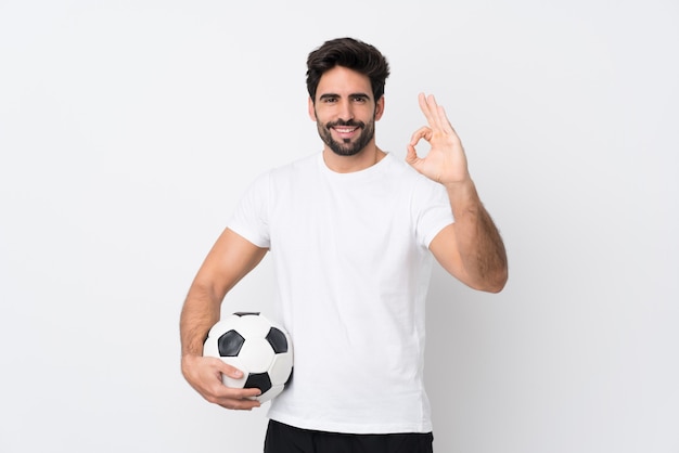 Jeune bel homme avec barbe sur mur blanc isolé avec ballon de football et faisant signe OK