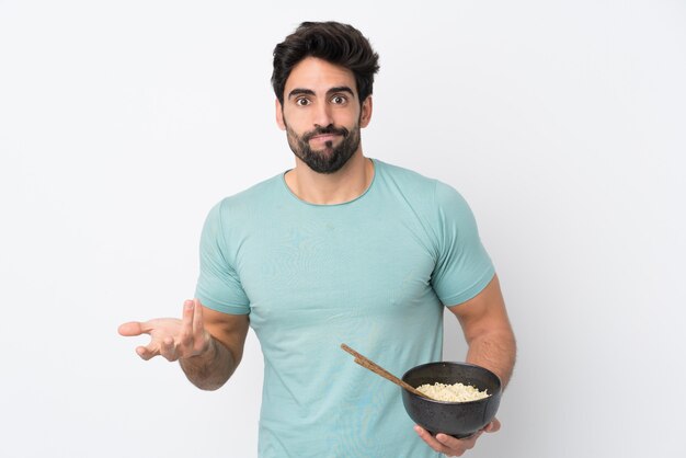 Jeune Bel Homme Avec Barbe Sur Mur Blanc Isolé Faisant Des Doutes Geste Tout En Soulevant Les épaules Tout En Tenant Un Bol De Nouilles Avec Des Baguettes