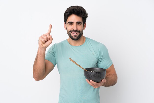 Jeune bel homme avec barbe sur mur blanc isolé pointant vers le haut une excellente idée tout en tenant un bol de nouilles avec des baguettes