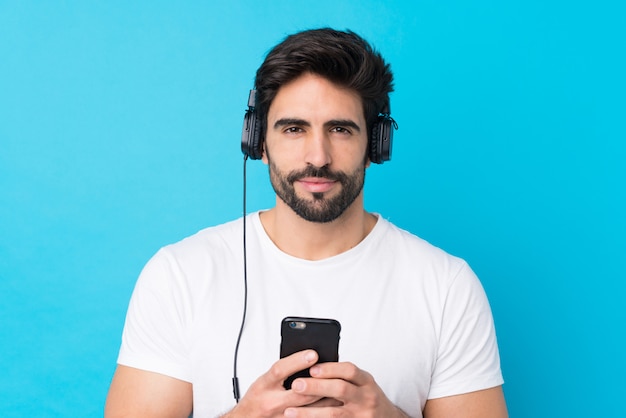 Jeune bel homme avec barbe sur mur bleu isolé écouter de la musique avec un mobile et à l'avant