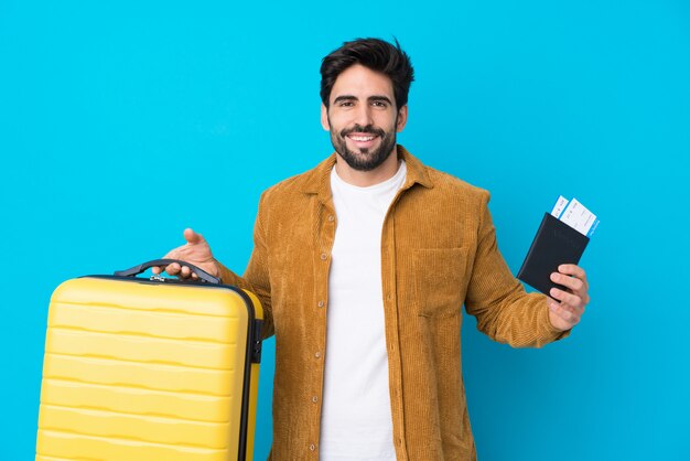 Jeune bel homme avec barbe sur mur bleu isolé en vacances avec valise et passeport