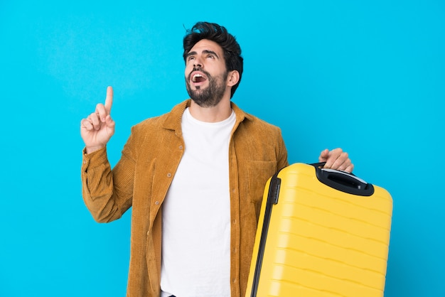 Jeune bel homme avec barbe sur mur bleu isolé en vacances avec valise de voyage et pointant vers le haut