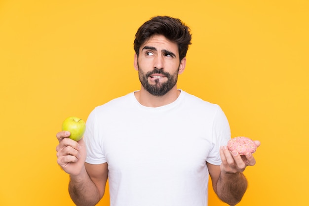 Jeune bel homme avec barbe sur mur jaune isolé ayant des doutes tout en prenant une tablette de chocolat dans une main et une pomme dans l'autre