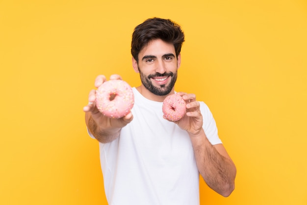 Jeune bel homme avec barbe sur mur jaune isolé tenant des beignets avec une expression heureuse