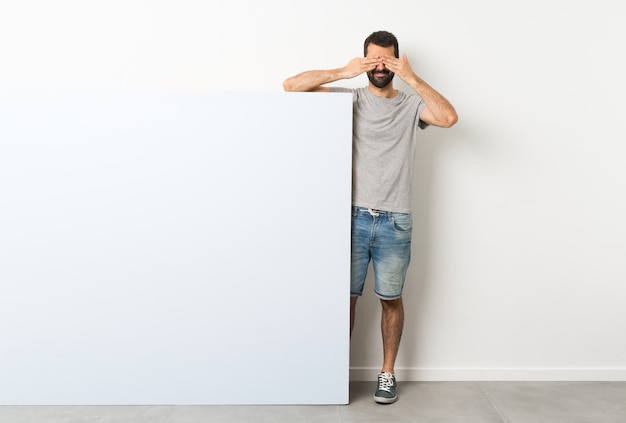 Jeune bel homme à la barbe tenant une grande pancarte vide bleue couvrant les yeux à la main