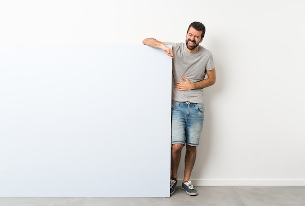 Jeune bel homme à la barbe tenant une grande pancarte vide bleue souriant beaucoup