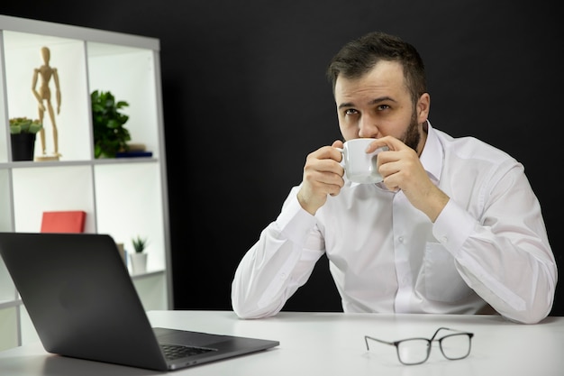 Jeune Bel Homme Barbu En Chemise Blanche Rêvant De Profiter D'une Tasse De Cappuccino, Pigiste Masculin Assis Au Bureau Devant Un Ordinateur Portable Dans Le Bureau à Domicile. Freelance, Travail à Distance, Concept D'indépendant