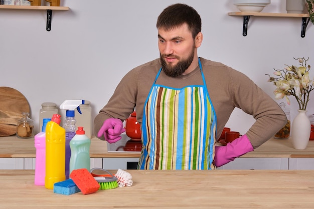 Jeune bel homme barbu dans la cuisine, montre tout son personnel de nettoyage - détergents, brosses, sprays. Il pense qu'il est prêt pour un vrai nettoyage