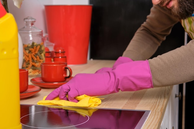 Jeune bel homme barbu dans la cuisine portant un tablier et des gants roses nettoie la surface de cuisson u...