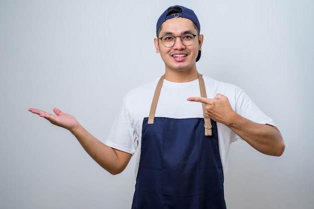 Jeune bel homme barista asiatique portant un tablier montrant le produit pointant vers quelque chose avec les mains