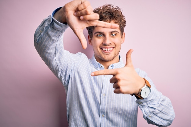 Jeune bel homme blond aux cheveux bouclés portant une chemise rayée sur fond blanc souriant faisant cadre avec les mains et les doigts avec un visage heureux Concept de créativité et de photographie