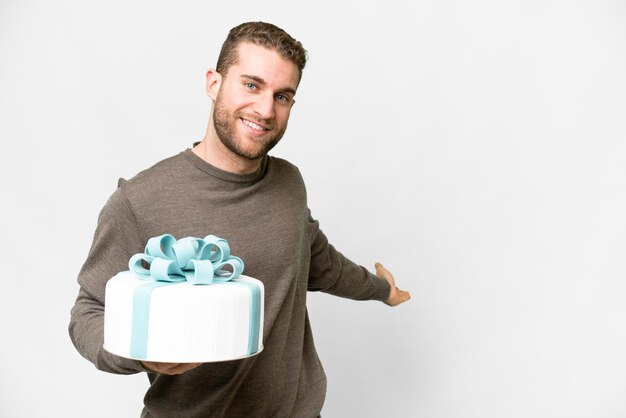 Jeune bel homme blond avec un gros gâteau sur fond blanc isolé tendant les mains sur le côté pour inviter à venir