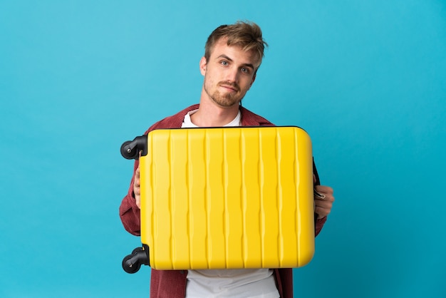 Jeune bel homme blond isolé sur bleu en vacances avec valise de voyage