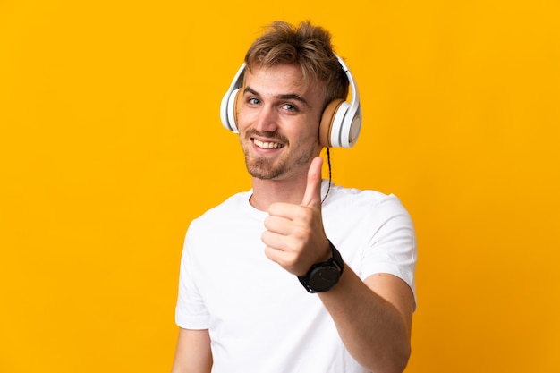 Jeune bel homme blond isolé sur un mur jaune à l'écoute de la musique et avec le pouce vers le haut