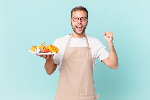 Jeune bel homme blond se sentant choqué, riant et célébrant le succès. concept de cuisson des gaufres