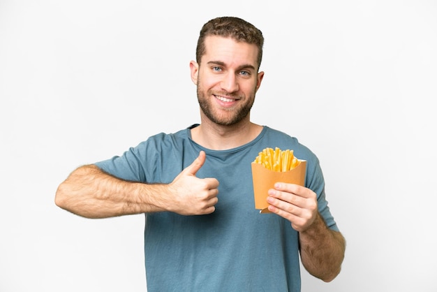 Jeune bel homme blond tenant des frites frites sur fond blanc isolé donnant un geste du pouce levé