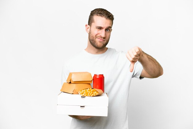 Jeune bel homme blond tenant des pizzas et des hamburgers sur fond isolé montrant le pouce vers le bas avec une expression négative