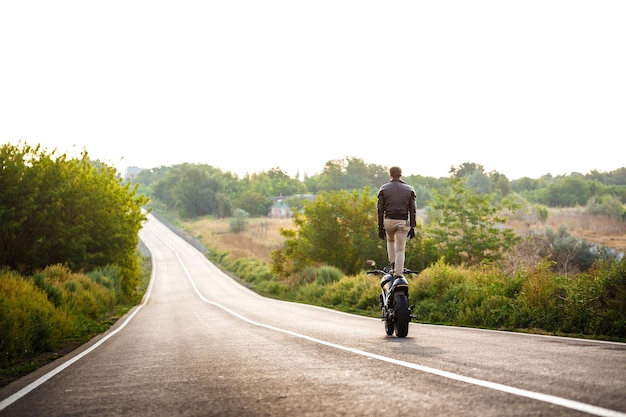 Jeune bel homme en blouson de cuir à moto, faisant des tours sur une route de campagne.