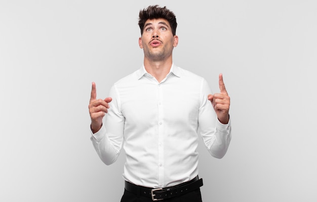 Jeune bel homme à la bouche choquée, étonnée et ouverte, pointant vers le haut avec les deux mains pour copier l'espace