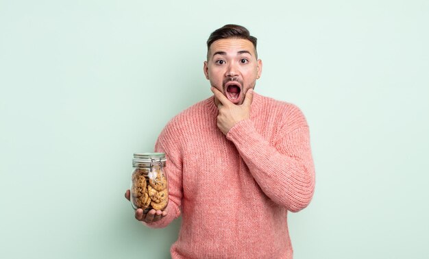 Jeune bel homme avec la bouche et les yeux grands ouverts et la main sur le menton. concept de bouteille de biscuits