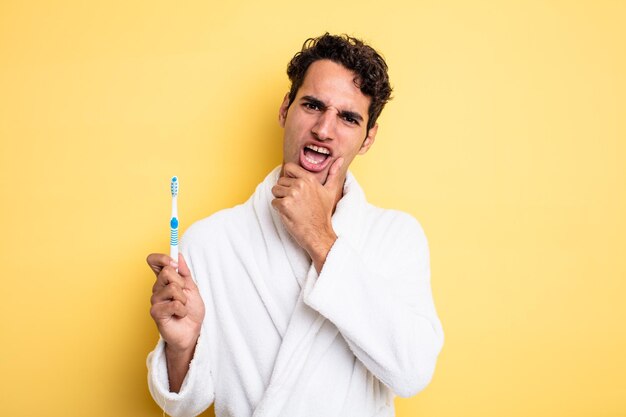 Jeune bel homme avec la bouche et les yeux grands ouverts et la main sur le menton. concept de peignoir et brosse à dents