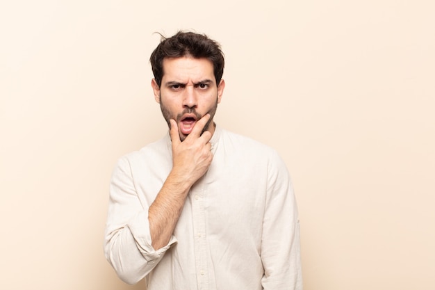 Jeune bel homme avec la bouche et les yeux grands ouverts et la main sur le menton, se sentant désagréablement choqué, disant quoi ou wow.