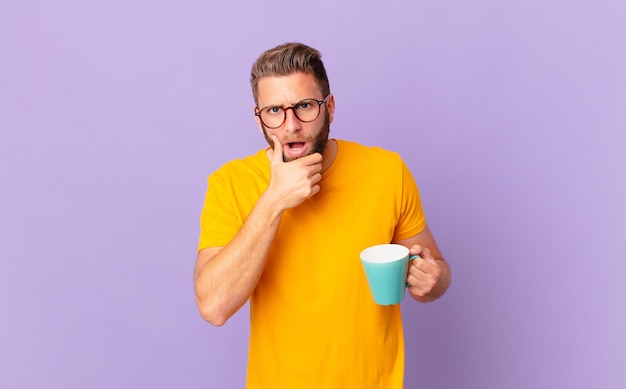 jeune bel homme avec la bouche et les yeux grands ouverts et la main sur le menton. et tenant une tasse de café