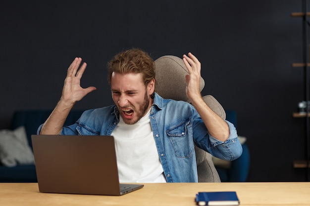 Jeune bel homme bouclé en colère à l'aide d'un ordinateur portable. Un pigiste stressé a perdu des données sur un ordinateur portable cassé qui ne fonctionnait pas. Recevoir de mauvaises nouvelles ou un e-mail, un message de spam. Les jeunes travaillant avec des appareils mobiles.