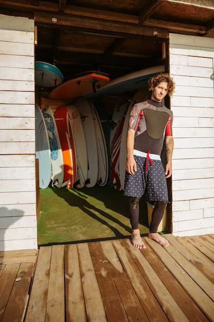 Jeune bel homme bouclé en maillot de bain debout à la cabane de surf à la plage