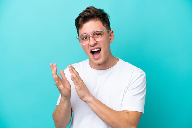 Jeune bel homme brésilien isolé sur fond bleu avec des lunettes et applaudissant