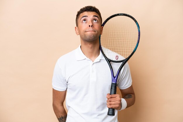 Jeune bel homme brésilien jouant au tennis isolé sur fond beige et levant les yeux