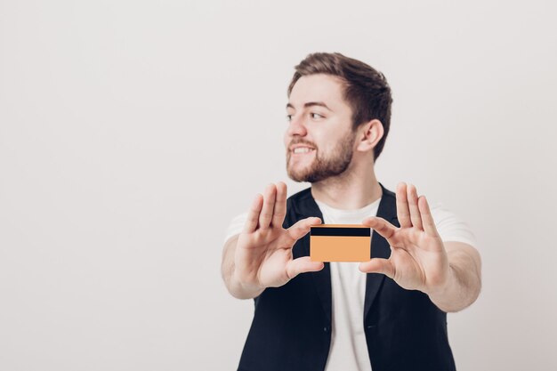 Jeune Bel Homme Brune Avec Une Barbe Dans Une Chemise Blanche Et Un Gilet Noir Tenant Une Carte De Crédit En Plastique Et Souriant. Se Concentrer Sur La Carte. Lumière Douce