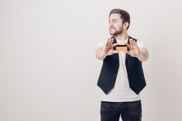 Jeune bel homme brune avec une barbe dans une chemise blanche et un gilet noir tenant une carte de crédit en plastique et souriant. se concentrer sur la carte. Lumière douce