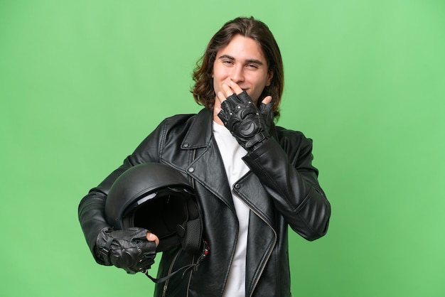 Jeune bel homme avec un casque de moto isolé sur fond vert chroma heureux et souriant couvrant la bouche avec la main