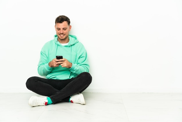 Jeune bel homme caucasien assis sur le sol, envoyer un message avec le mobile