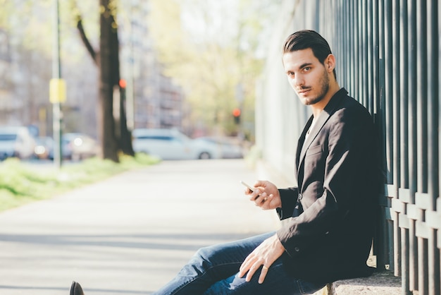 Jeune bel homme caucasien assis tenant un téléphone intelligent, regardant à huis clos, souriant