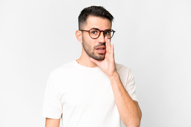 Jeune bel homme caucasien sur fond blanc isolé chuchotant quelque chose avec un geste de surprise tout en regardant sur le côté