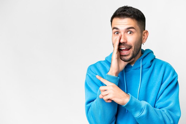 Jeune bel homme caucasien sur fond blanc isolé pointant vers le côté pour présenter un produit et chuchotant quelque chose