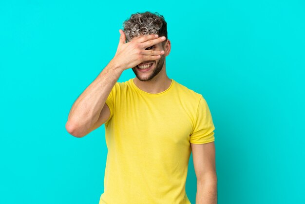 Jeune bel homme caucasien isolé sur fond bleu couvrant les yeux par les mains et souriant