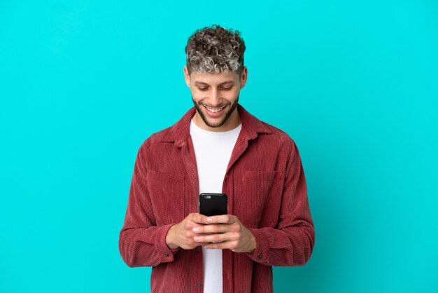 Jeune bel homme caucasien isolé sur fond bleu envoyant un message ou un e-mail avec le mobile