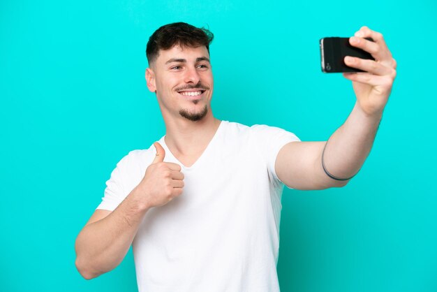 Jeune bel homme caucasien isolé sur fond bleu faisant un selfie avec un téléphone portable