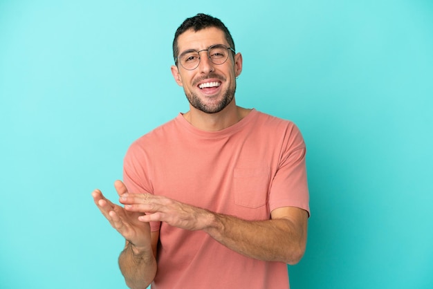 Jeune bel homme caucasien isolé sur fond bleu avec des lunettes et applaudissant